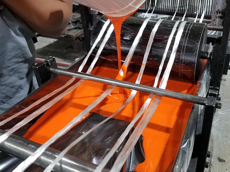 Image of industrial machinery. Technician is pouring orange pigment to a resin bath. Fiberglass tow is threaded through the machinery, where it will be dipped in the colored resin in preparation for filament winding.